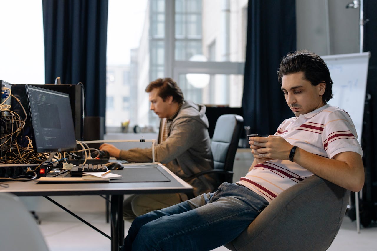 about-01 Two men working in a modern office, one on a computer and the other on a smartphone.