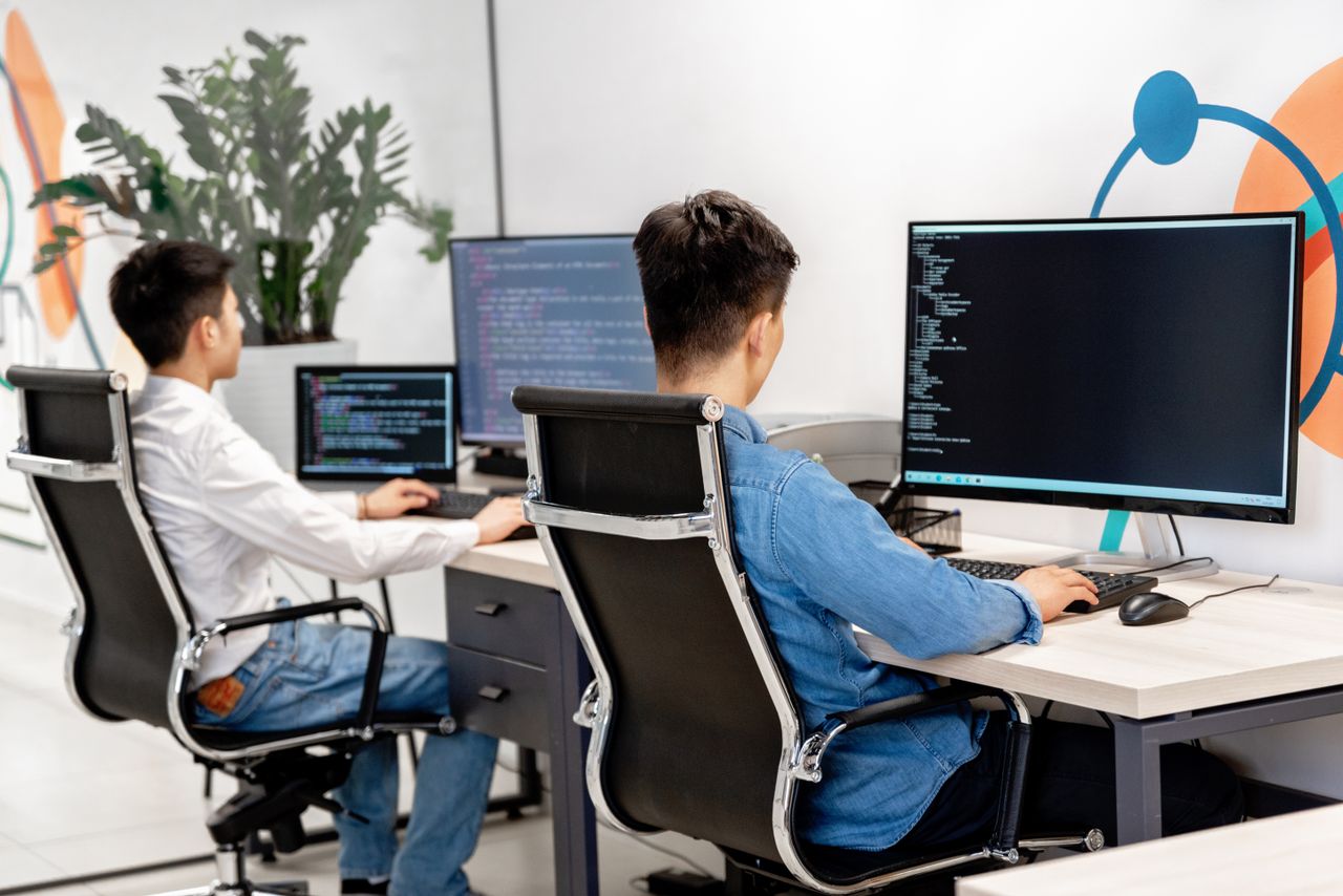 hero-img-02 Two male developers at desks programming in a modern office workspace with large monitors.